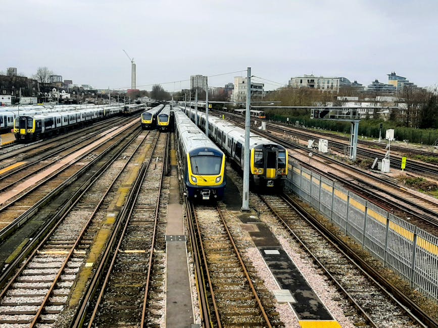 A busy train yard viewed from an elevated perspective, showcasing multiple parallel railway tracks with some used for passenger trains and others empty. Two modern, yellow and grey electric trains are